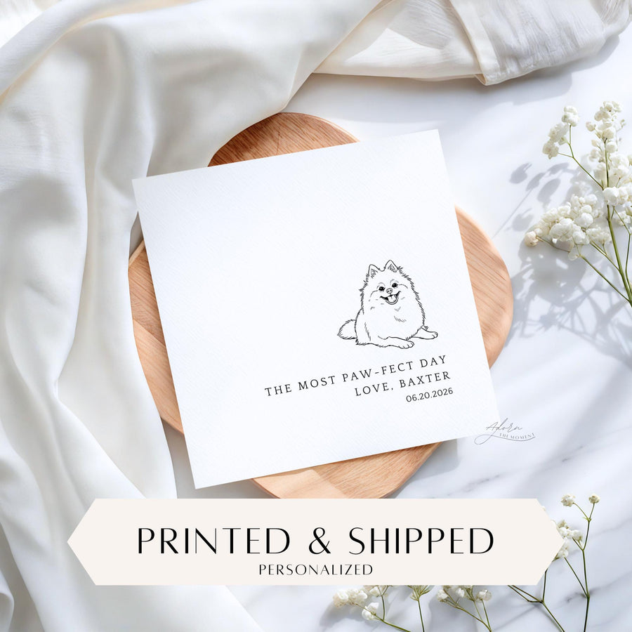 May include: A white card with a black line drawing of a smiling dog. The text reads "THE MOST PAW-FECT DAY LOVE, BAXTER 06.20.2026". The card is on a wooden tray with white fabric and flowers in the background.
