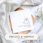 May include: A white card with a black line drawing of a smiling dog. The text reads "THE MOST PAW-FECT DAY LOVE, BAXTER 06.20.2026". The card is on a wooden tray with white fabric and flowers in the background.
